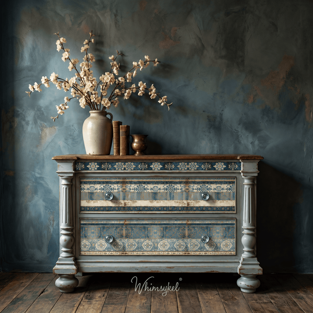 A distressed blue and beige wooden chest of drawers styled with a ceramic vase of cherry blossoms, books, and a small brass candle holder, set against a textured dark blue wall. Featuring 3 Papered Past Designs
